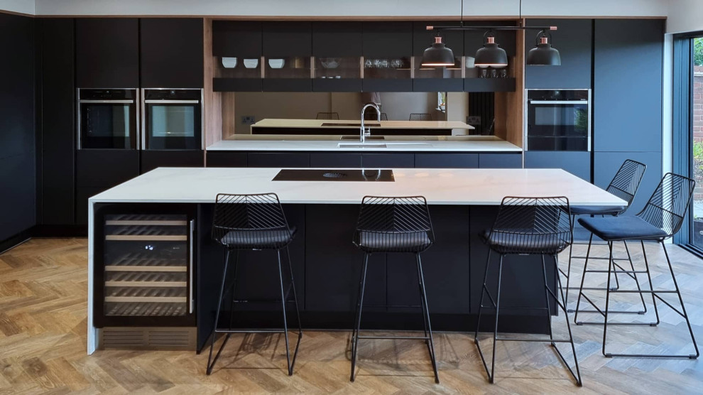 This is an example of a large modern single-wall open plan kitchen in Hertfordshire with an integrated sink, flat-panel cabinets, black cabinets, quartz worktops, mirror splashback, black appliances, light hardwood flooring, an island, multi-coloured floors, white worktops and feature lighting.