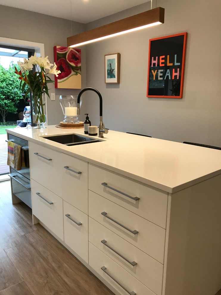 This is an example of a large scandinavian galley separate kitchen in Christchurch with a double-bowl sink, flat-panel cabinets, white cabinets, quartz benchtops, grey splashback, porcelain splashback, stainless steel appliances, vinyl floors, with island and brown floor.