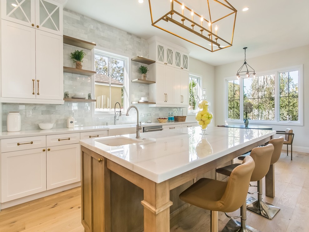Design ideas for a transitional galley eat-in kitchen in Los Angeles with a farmhouse sink, shaker cabinets, white cabinets, white splashback, subway tile splashback, light hardwood floors, with island and beige floor.