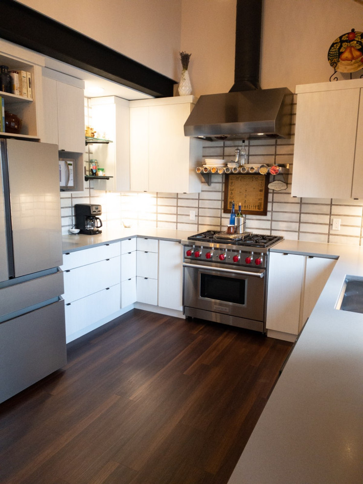 Foto på ett funkis grå kök, med en undermonterad diskho, släta luckor, vita skåp, bänkskiva i kvarts, vitt stänkskydd, stänkskydd i porslinskakel, rostfria vitvaror, mellanmörkt trägolv, en köksö och brunt golv