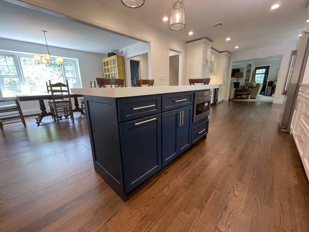 Photo of a large traditional l-shaped kitchen/diner in New York with a submerged sink, recessed-panel cabinets, blue cabinets, engineered stone countertops, white splashback, ceramic splashback, stainless steel appliances, medium hardwood flooring, an island, brown floors and white worktops.