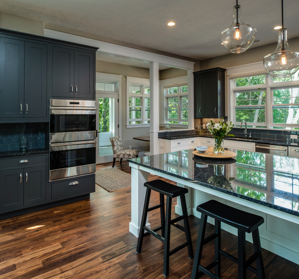 Large transitional single-wall medium tone wood floor open concept kitchen photo with an undermount sink, shaker cabinets, gray cabinets, granite countertops, gray backsplash, ceramic backsplash, stainless steel appliances and an island