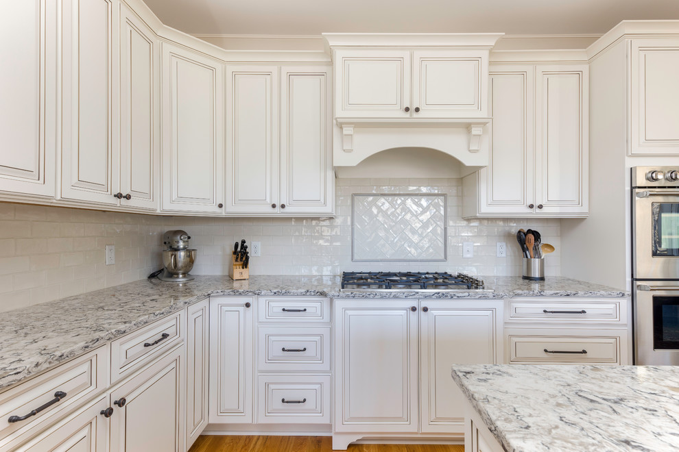 This is an example of a mid-sized traditional l-shaped kitchen in Nashville with raised-panel cabinets, white cabinets, quartz benchtops, white splashback, ceramic splashback, stainless steel appliances, medium hardwood floors, with island, brown floor and multi-coloured benchtop.