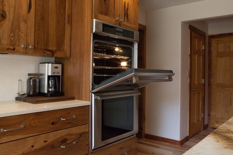 Example of a mid-sized mountain style l-shaped light wood floor eat-in kitchen design in Indianapolis with a single-bowl sink, flat-panel cabinets, medium tone wood cabinets, quartz countertops, white backsplash, subway tile backsplash, stainless steel appliances, white walls, an island and beige countertops