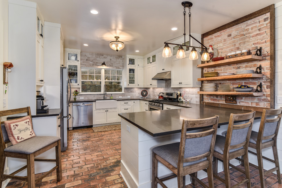 Photo of a large country u-shaped kitchen in Other with a farmhouse sink, shaker cabinets, white cabinets, brick splashback, stainless steel appliances, brick floors, a peninsula, red floor and beige splashback.