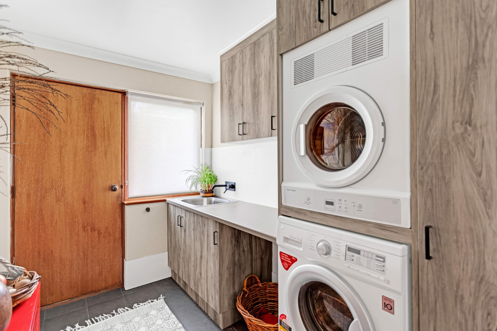 Small transitional single-wall dedicated laundry room in Adelaide with a drop-in sink, flat-panel cabinets, light wood cabinets, laminate benchtops, white splashback, porcelain splashback, white walls, porcelain floors, a stacked washer and dryer, grey floor and grey benchtop.