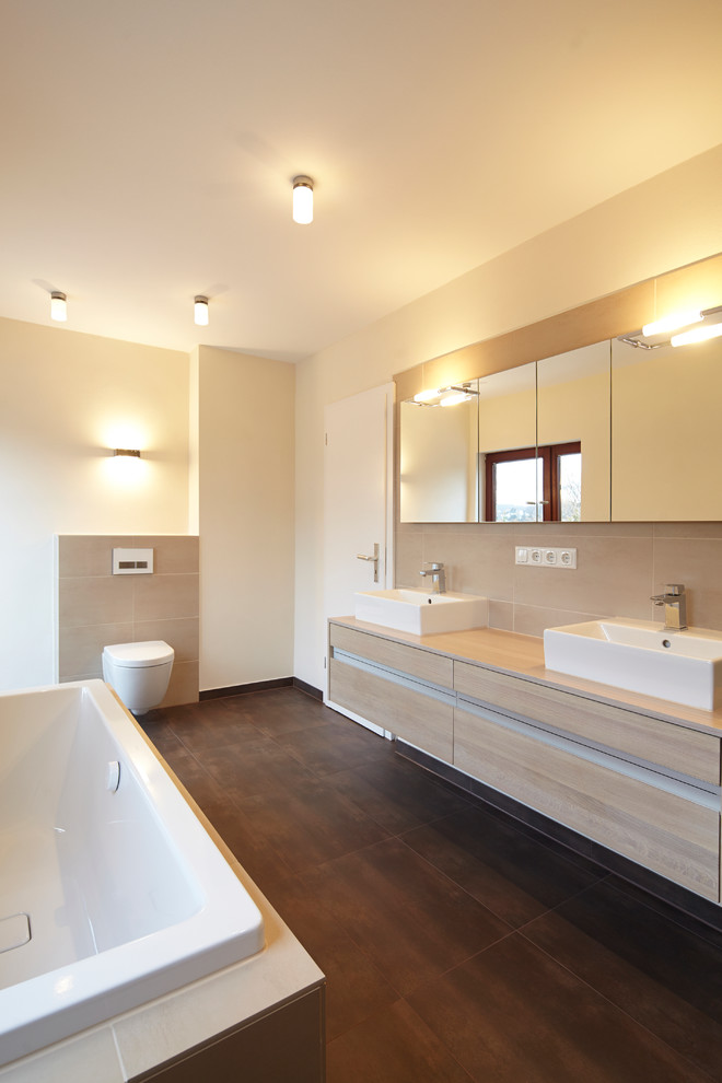 Large contemporary bathroom in Dortmund with a vessel sink, beige tile, flat-panel cabinets, medium wood cabinets, a drop-in tub, a wall-mount toilet, beige walls, wood benchtops and beige benchtops.