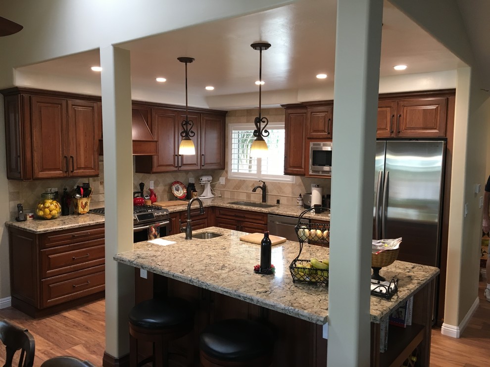 Large traditional l-shaped open plan kitchen in Orange County with an undermount sink, raised-panel cabinets, medium wood cabinets, granite benchtops, beige splashback, ceramic splashback, stainless steel appliances, medium hardwood floors, with island and brown floor.