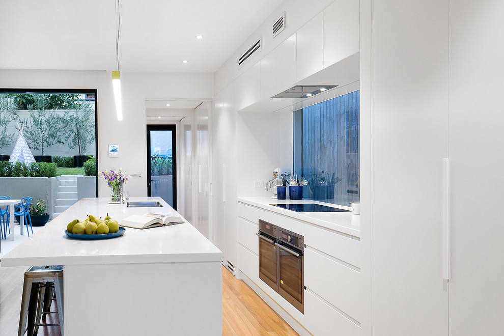 Contemporary galley kitchen in Sydney with flat-panel cabinets, white cabinets, window splashback, stainless steel appliances, medium hardwood floors, with island, brown floor, white benchtop and an undermount sink.