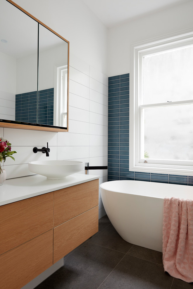 Photo of a large modern master bathroom in Melbourne with glass-front cabinets, medium wood cabinets, a freestanding tub, white tile, ceramic tile, white walls, porcelain floors, a vessel sink, black floor and white benchtops.