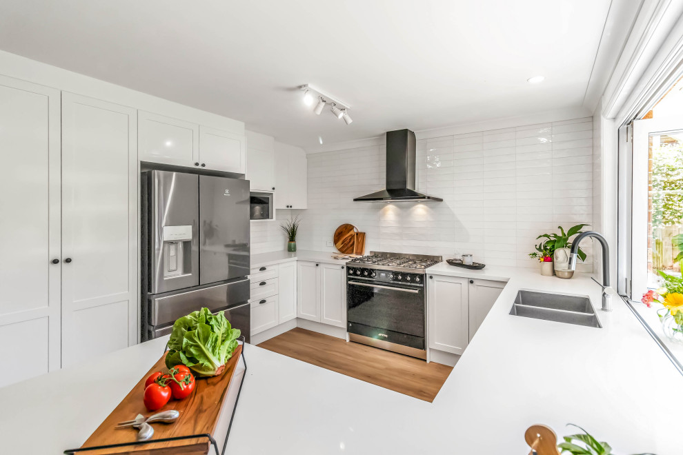 Design ideas for a mid-sized traditional u-shaped open plan kitchen in Sydney with an undermount sink, shaker cabinets, white cabinets, quartz benchtops, white splashback, subway tile splashback, stainless steel appliances, vinyl floors, a peninsula, brown floor and white benchtop.