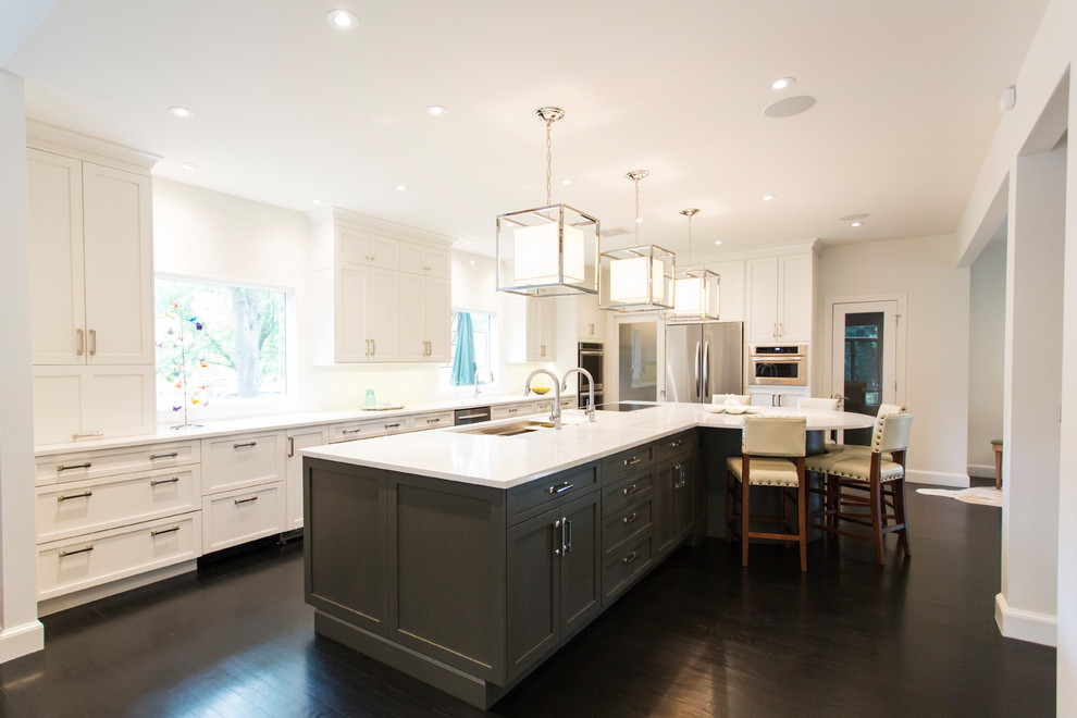 Large transitional l-shaped dark wood floor and brown floor kitchen photo with an undermount sink, shaker cabinets, white cabinets, quartz countertops, white backsplash, stainless steel appliances, an island and white countertops