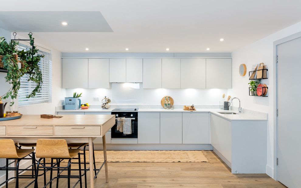 This is an example of a scandinavian l-shaped eat-in kitchen in Dorset with an undermount sink, flat-panel cabinets, white cabinets, white splashback, black appliances, light hardwood floors, no island, beige floor and white benchtop.