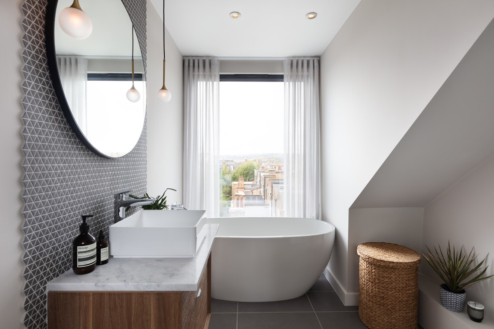 This is an example of a contemporary bathroom in London with flat-panel cabinets, medium wood cabinets, a freestanding tub, grey walls, a vessel sink, grey floor and white benchtops.