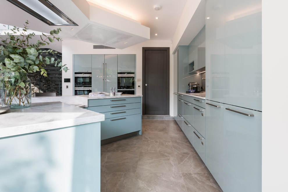 Mid-sized modern l-shaped eat-in kitchen in Gloucestershire with a drop-in sink, flat-panel cabinets, blue cabinets, solid surface benchtops, metallic splashback, glass sheet splashback, black appliances, ceramic floors, with island, brown floor and beige benchtop.