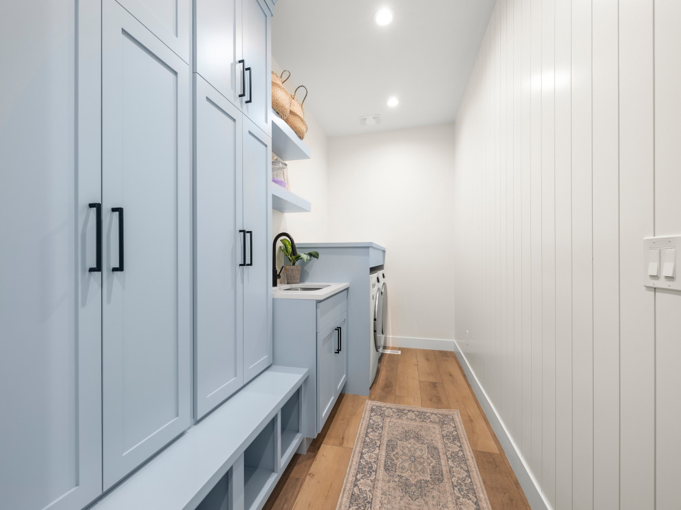 Design ideas for a large transitional single-wall dedicated laundry room in Other with an undermount sink, shaker cabinets, blue cabinets, quartz benchtops, white walls, light hardwood floors, a side-by-side washer and dryer, brown floor, white benchtop and planked wall panelling.