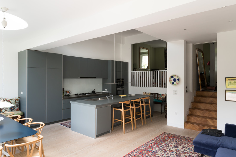 This is an example of a mid-sized contemporary u-shaped open plan kitchen in London with an integrated sink, flat-panel cabinets, grey cabinets, solid surface benchtops, blue splashback, glass sheet splashback, stainless steel appliances, light hardwood floors, with island and beige floor.