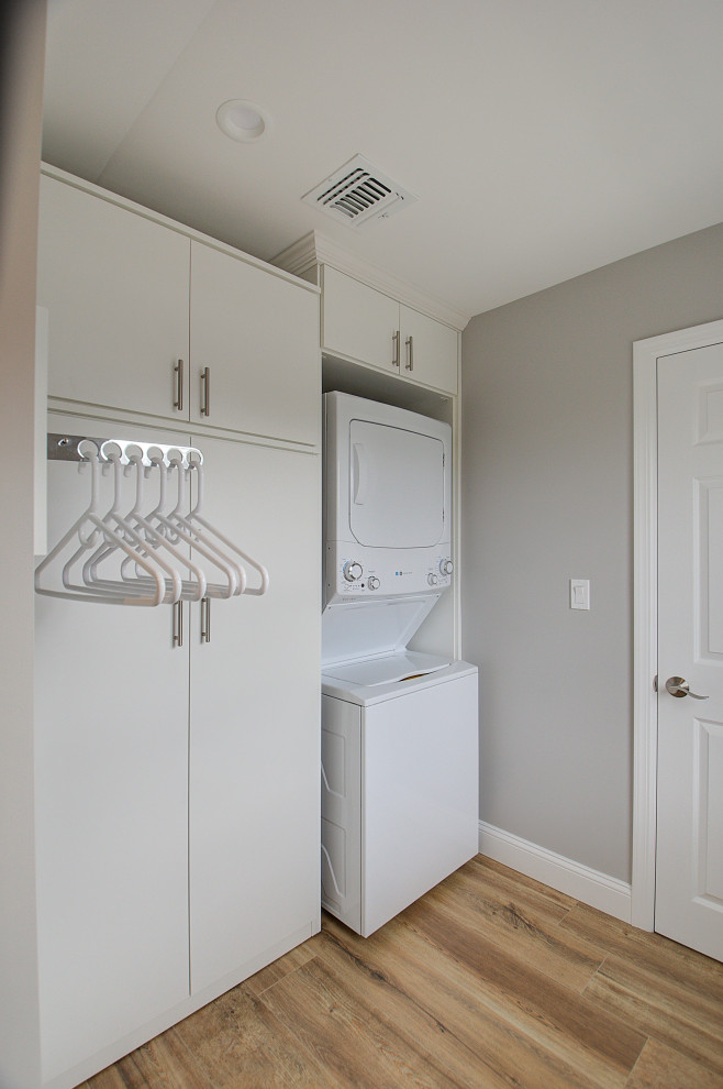 Photo of a mid-sized transitional galley dedicated laundry room in New York with flat-panel cabinets, white cabinets, grey walls, porcelain floors, a stacked washer and dryer and beige floor.