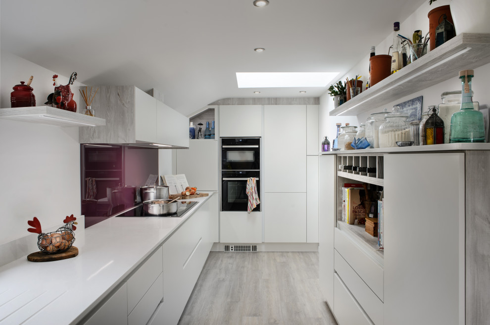 Small contemporary u-shaped separate kitchen in Cornwall with a drop-in sink, flat-panel cabinets, white cabinets, onyx benchtops, red splashback, glass sheet splashback, black appliances, light hardwood floors, no island, beige floor and white benchtop.