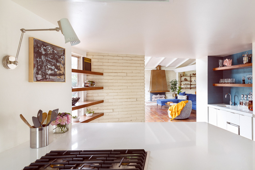 This is an example of a large midcentury galley kitchen in Houston with a double-bowl sink, flat-panel cabinets, white cabinets, solid surface benchtops, multi-coloured splashback, mosaic tile splashback, stainless steel appliances, terra-cotta floors, with island, brown floor and white benchtop.