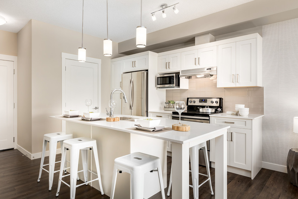 Photo of a mid-sized transitional galley eat-in kitchen in Calgary with an undermount sink, shaker cabinets, white cabinets, quartz benchtops, beige splashback, ceramic splashback, stainless steel appliances, dark hardwood floors, with island, brown floor and white benchtop.