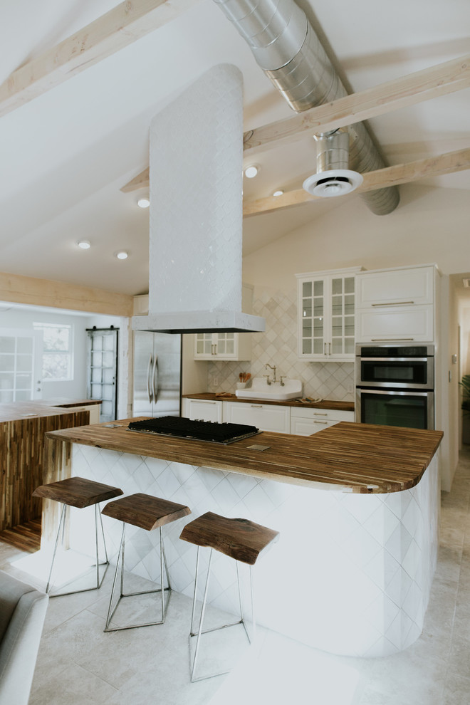 Design ideas for a mid-sized scandinavian u-shaped open plan kitchen in Phoenix with a farmhouse sink, glass-front cabinets, white cabinets, wood benchtops, white splashback, ceramic splashback, stainless steel appliances, porcelain floors, multiple islands, grey floor and brown benchtop.