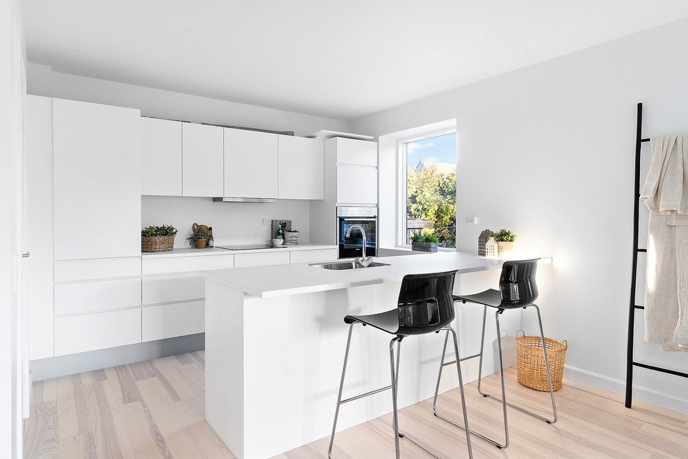 Scandinavian kitchen in Copenhagen with a single-bowl sink, flat-panel cabinets, white cabinets, stainless steel appliances, light hardwood floors, a peninsula and white benchtop.