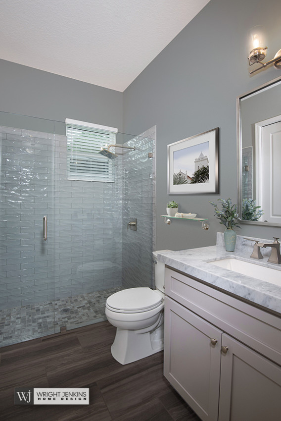 Design ideas for a mid-sized transitional 3/4 bathroom in Orlando with flat-panel cabinets, grey cabinets, a curbless shower, a one-piece toilet, blue tile, glass tile, blue walls, medium hardwood floors, an undermount sink, marble benchtops, brown floor, a hinged shower door and white benchtops.