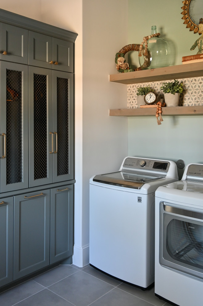 This is an example of a mid-sized l-shaped utility room with a farmhouse sink, recessed-panel cabinets, green cabinets, wood benchtops, multi-coloured splashback, mosaic tile splashback, green walls, porcelain floors, a side-by-side washer and dryer, grey floor and grey benchtop.