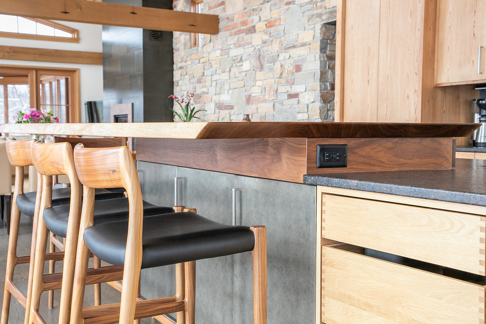 Example of a large mountain style l-shaped medium tone wood floor and brown floor open concept kitchen design with flat-panel cabinets, light wood cabinets, granite countertops, black backsplash, stainless steel appliances, an island, black countertops, a double-bowl sink and porcelain backsplash