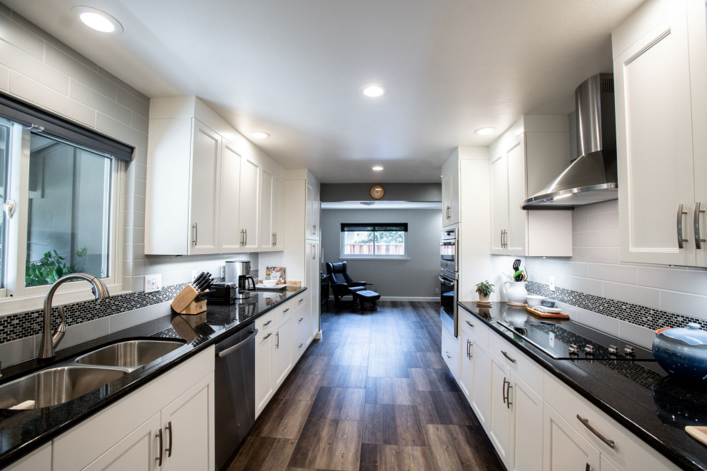 This is an example of a small contemporary galley kitchen in San Francisco with a double-bowl sink, recessed-panel cabinets, white cabinets, grey splashback, subway tile splashback, stainless steel appliances, no island, brown floor and black benchtop.