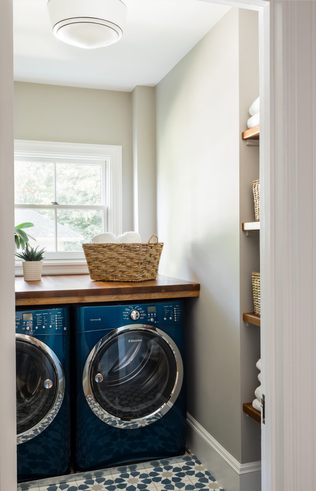 Inredning av en klassisk mellanstor bruna linjär brunt tvättstuga enbart för tvätt, med öppna hyllor, bruna skåp, träbänkskiva, grå väggar, klinkergolv i keramik, en tvättmaskin och torktumlare bredvid varandra och blått golv
