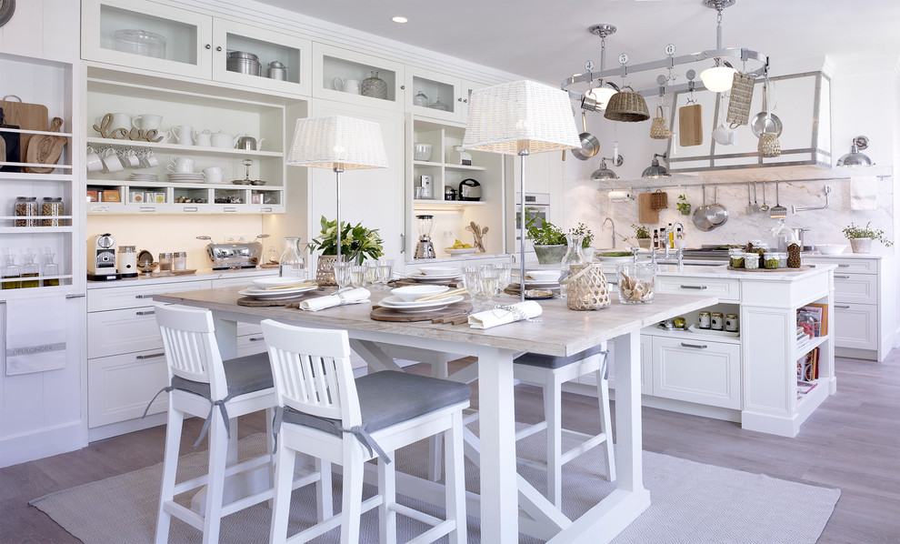 This is an example of a large country l-shaped open plan kitchen in Barcelona with an undermount sink, white cabinets, marble benchtops, white splashback, stone slab splashback, white appliances, light hardwood floors, with island and recessed-panel cabinets.