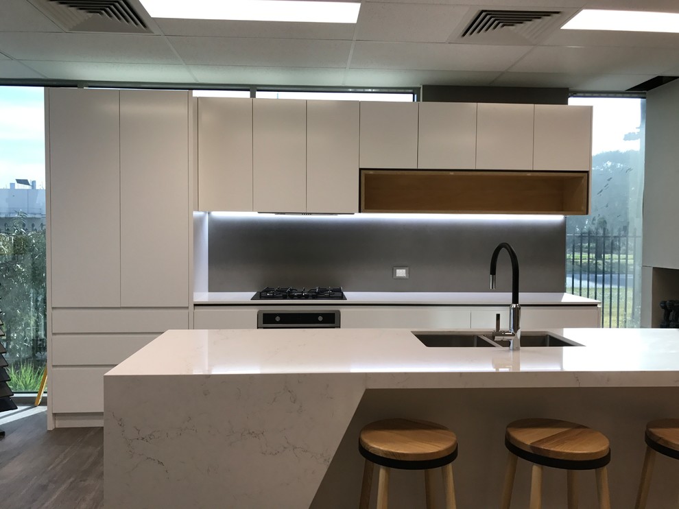 This is an example of a mid-sized modern galley open plan kitchen in Melbourne with an undermount sink, flat-panel cabinets, white cabinets, quartz benchtops, grey splashback, porcelain splashback, black appliances, vinyl floors, with island and grey floor.