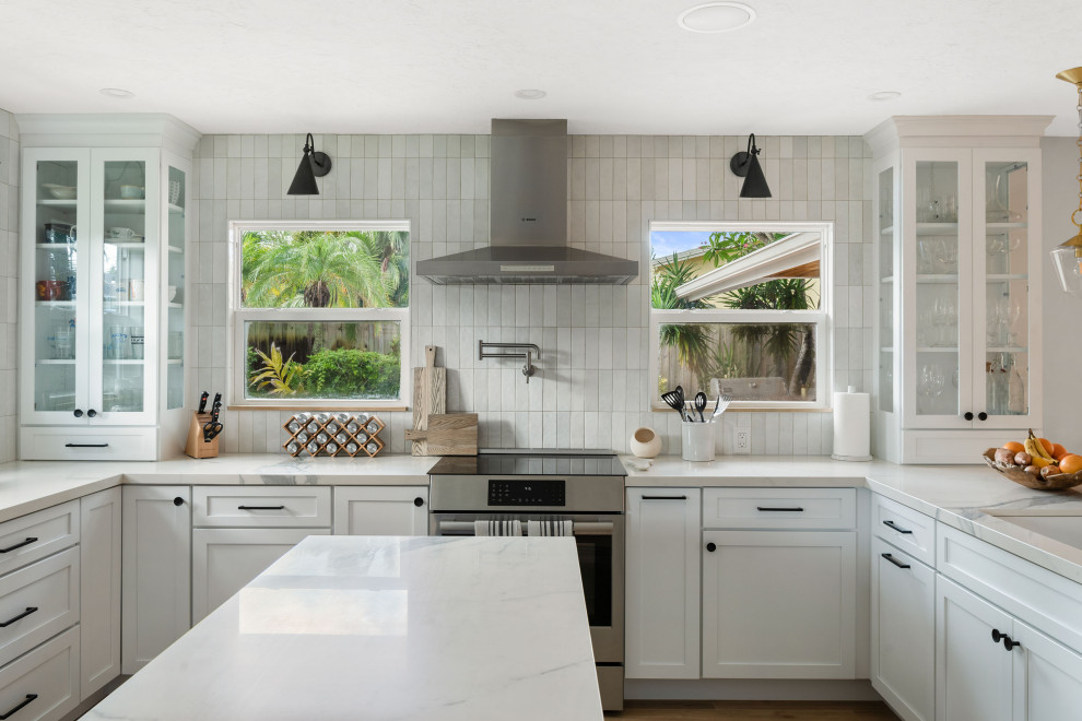 Photo of a medium sized beach style u-shaped kitchen/diner in Tampa with a submerged sink, shaker cabinets, white cabinets, engineered stone countertops, white splashback, porcelain splashback, stainless steel appliances, medium hardwood flooring, an island, brown floors and white worktops.