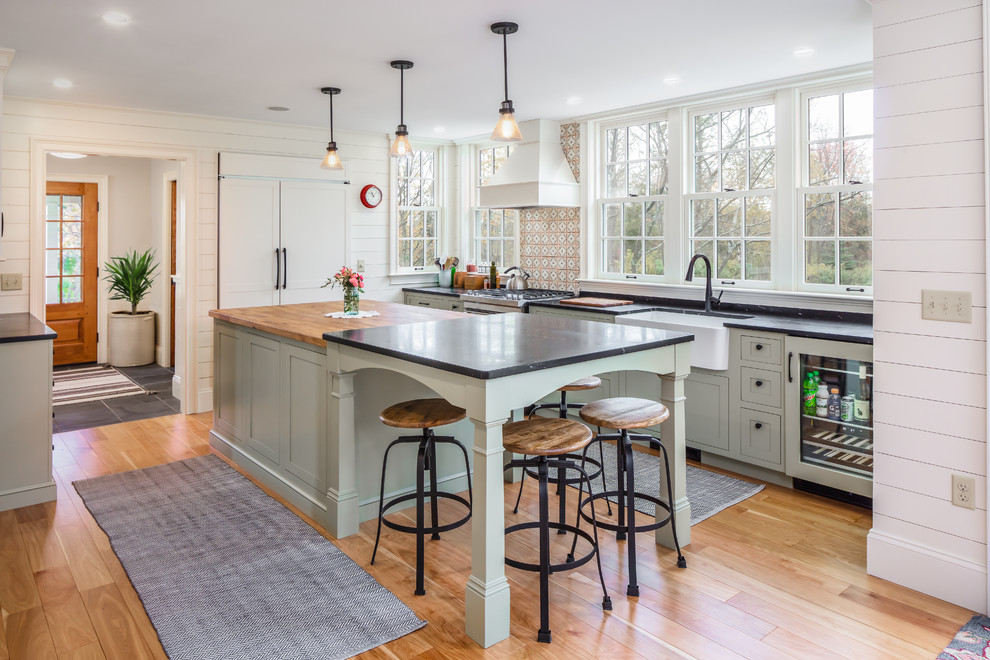 L-shaped medium tone wood floor and brown floor kitchen photo in Burlington with a farmhouse sink, shaker cabinets, gray cabinets, multicolored backsplash, paneled appliances, an island and black countertops