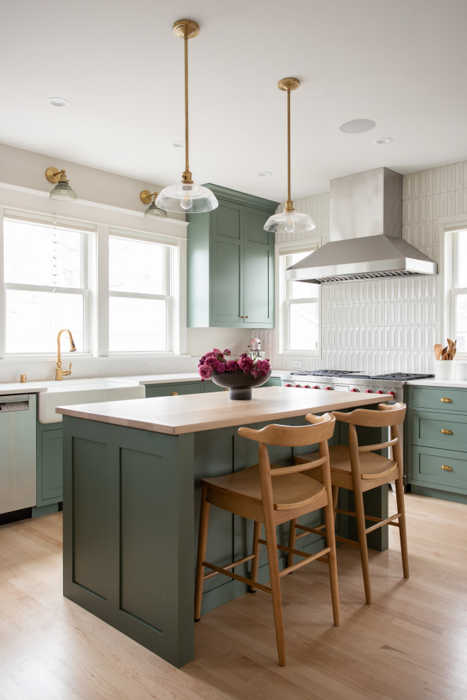 Design ideas for a medium sized traditional l-shaped kitchen/diner in Minneapolis with a belfast sink, recessed-panel cabinets, green cabinets, white splashback, ceramic splashback, stainless steel appliances, light hardwood flooring, an island, brown floors, wood worktops and brown worktops.