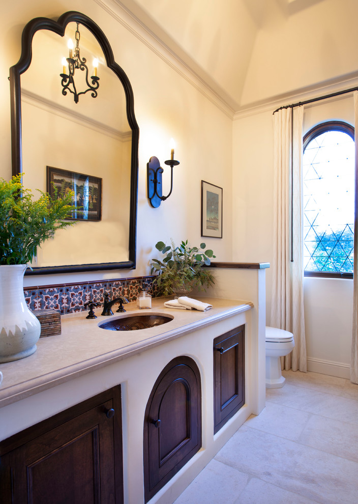 This is an example of a mediterranean 3/4 bathroom in San Diego with recessed-panel cabinets, brown cabinets, a one-piece toilet, terra-cotta tile, white walls, limestone floors, an undermount sink, limestone benchtops, beige floor and beige benchtops.