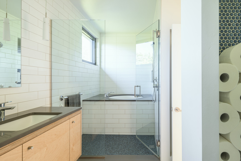 На фото: главная ванная комната среднего размера в современном стиле с фасадами с утопленной филенкой, светлыми деревянными фасадами, ванной в нише, душем над ванной, белой плиткой, керамической плиткой, белыми стенами, полом из керамической плитки, врезной раковиной, столешницей из искусственного камня, синим полом, душем с распашными дверями, серой столешницей, нишей, тумбой под две раковины и подвесной тумбой