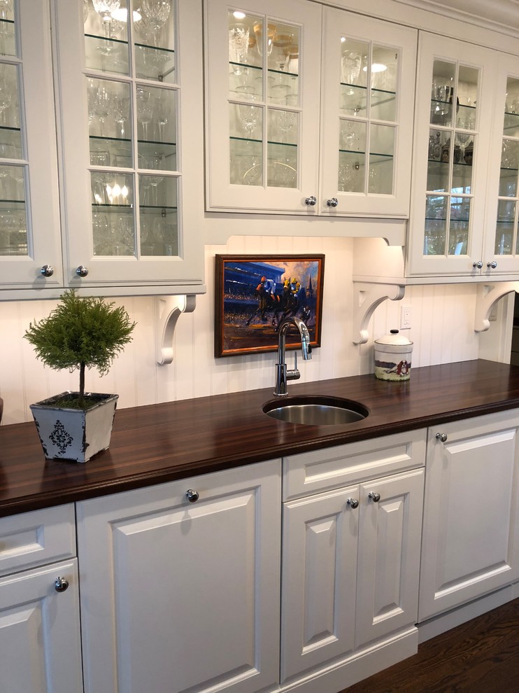 This is an example of a mid-sized traditional u-shaped wet bar in Philadelphia with an undermount sink, glass-front cabinets, white cabinets, wood benchtops, white splashback, timber splashback, medium hardwood floors and brown benchtop.