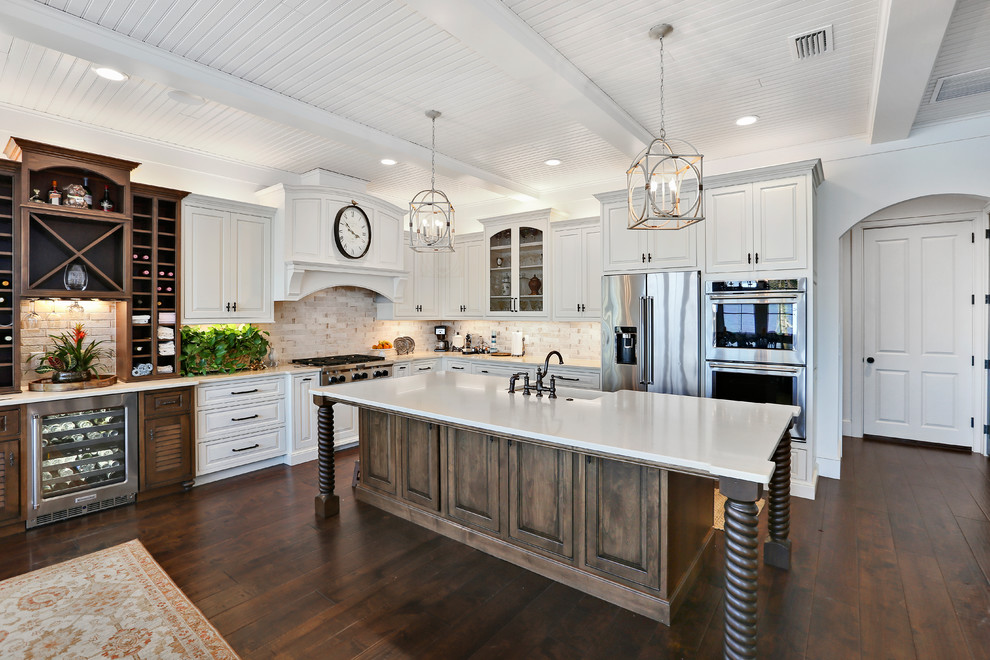 This is an example of a tropical l-shaped kitchen in Orlando with a farmhouse sink, raised-panel cabinets, white cabinets, beige splashback, stainless steel appliances, dark hardwood floors, with island, brown floor and white benchtop.