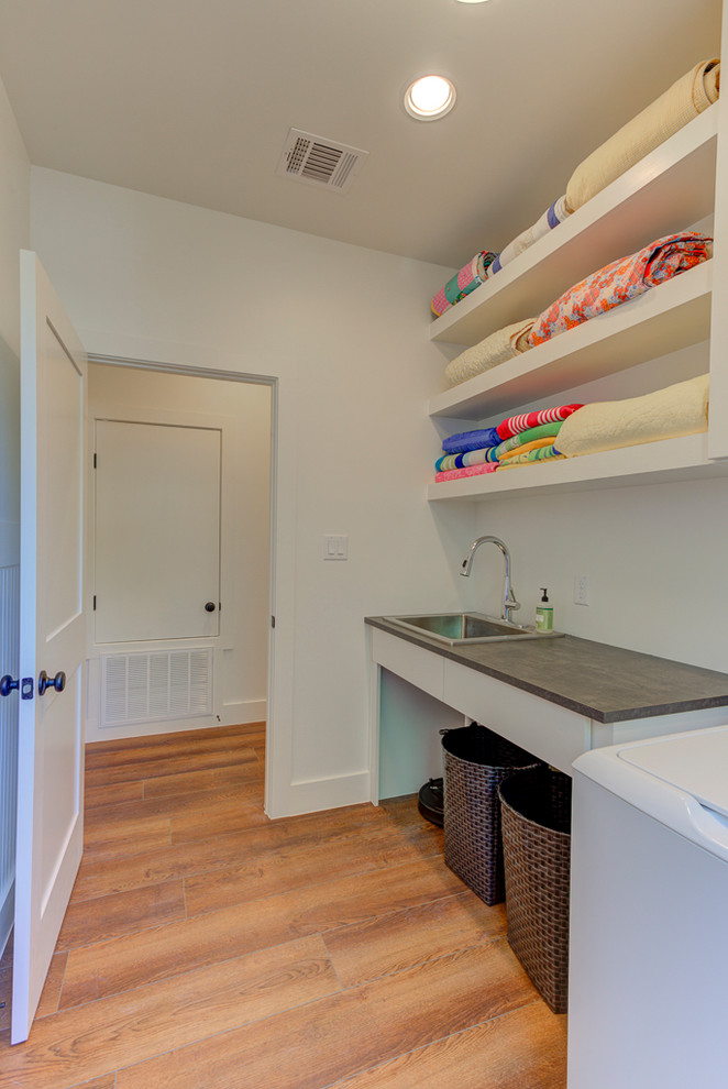 Photo of a small country galley dedicated laundry room in Austin with a drop-in sink, glass-front cabinets, grey cabinets, solid surface benchtops, white walls, vinyl floors, a side-by-side washer and dryer, brown floor and black benchtop.