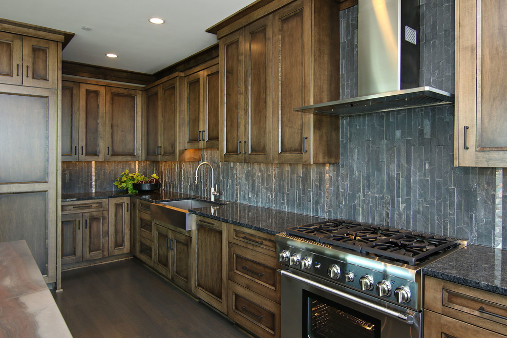 Photo of a large industrial l-shaped eat-in kitchen in Minneapolis with a farmhouse sink, recessed-panel cabinets, dark wood cabinets, granite benchtops, black splashback, stone tile splashback, stainless steel appliances, dark hardwood floors and with island.