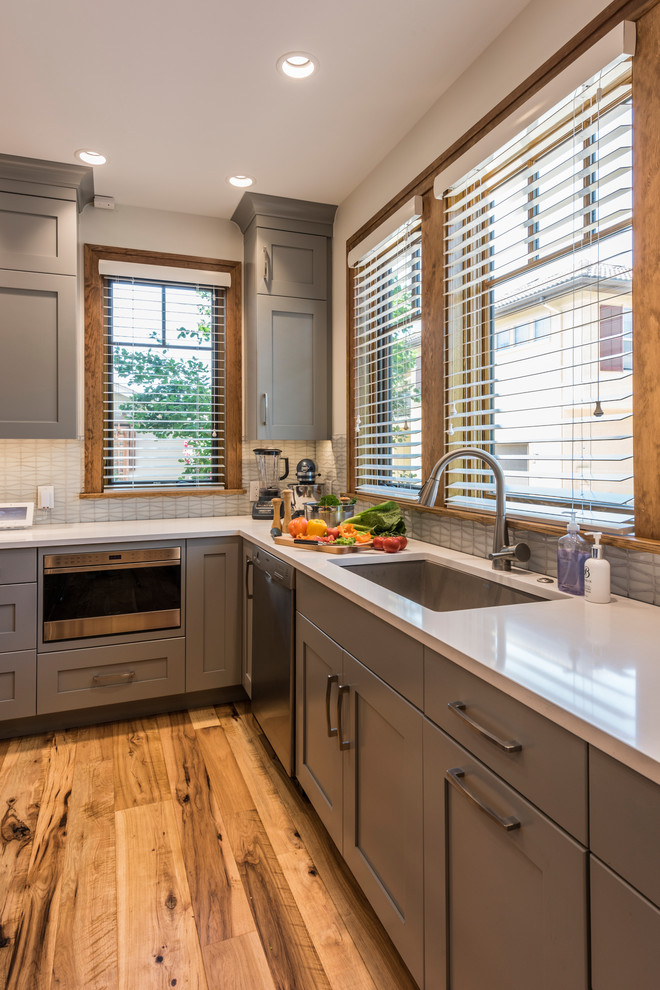 Mid-sized transitional l-shaped light wood floor and yellow floor eat-in kitchen photo in Denver with an undermount sink, shaker cabinets, gray cabinets, quartz countertops, white backsplash, glass tile backsplash, stainless steel appliances, an island and white countertops