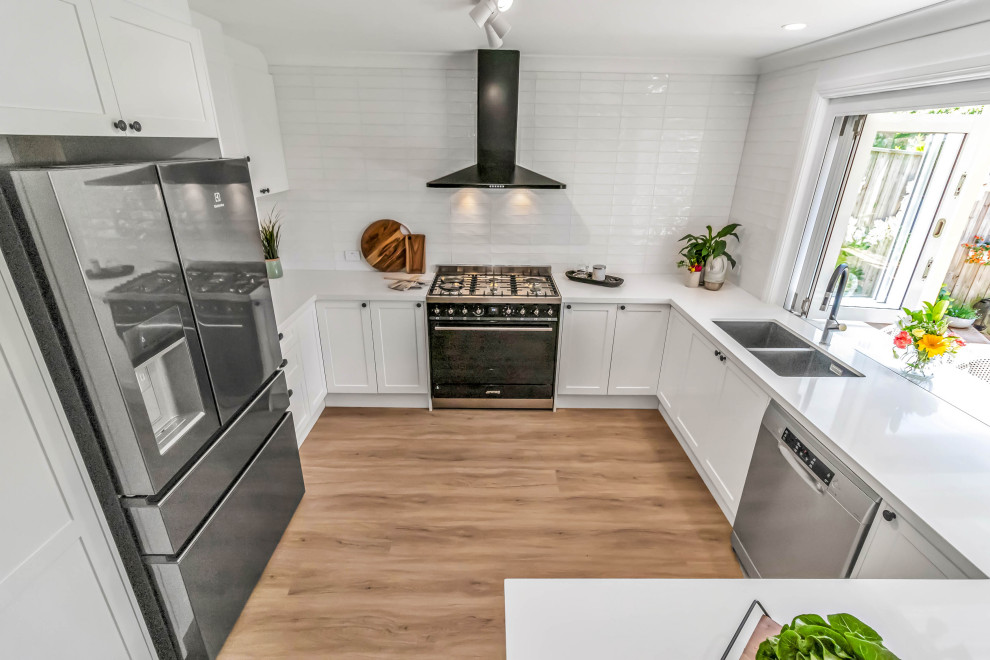 This is an example of a mid-sized traditional u-shaped open plan kitchen in Sydney with an undermount sink, shaker cabinets, white cabinets, quartz benchtops, white splashback, subway tile splashback, stainless steel appliances, vinyl floors, a peninsula, brown floor and white benchtop.