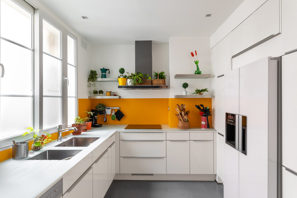 This is an example of a mid-sized contemporary l-shaped kitchen in Paris with a double-bowl sink, flat-panel cabinets, white cabinets, glass benchtops, panelled appliances, ceramic floors, no island, grey floor, white benchtop, orange splashback and glass sheet splashback.