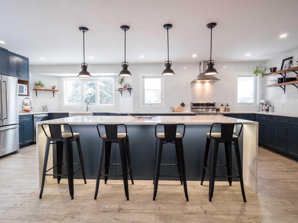 Design ideas for a large contemporary u-shaped open plan kitchen in Edmonton with a farmhouse sink, shaker cabinets, blue cabinets, white splashback, stainless steel appliances, with island, marble benchtops, subway tile splashback, medium hardwood floors, brown floor and white benchtop.
