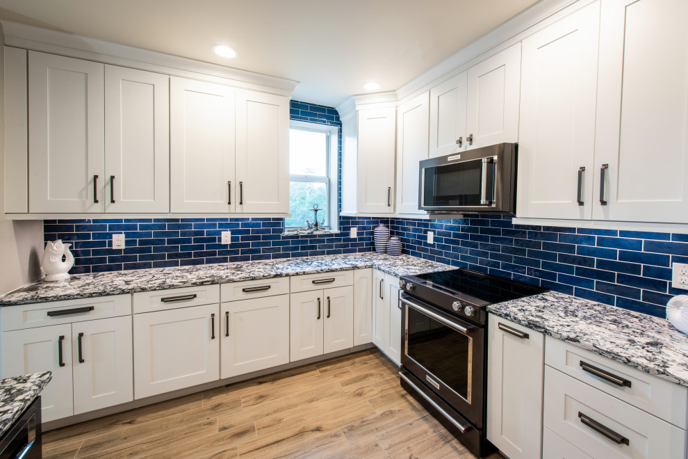 This is an example of a mid-sized beach style l-shaped eat-in kitchen in Orlando with an undermount sink, shaker cabinets, white cabinets, quartz benchtops, blue splashback, ceramic splashback, black appliances, porcelain floors, a peninsula, beige floor and blue benchtop.