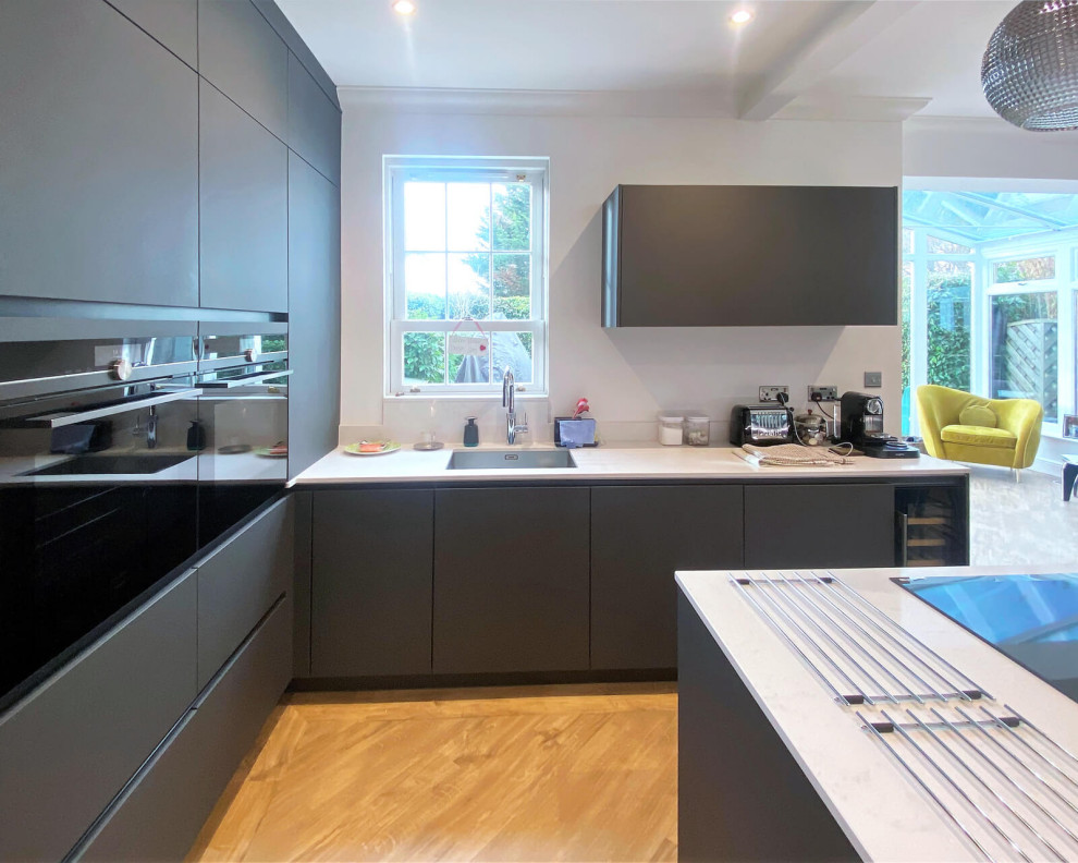 Design ideas for a small modern l-shaped open plan kitchen in London with a single-bowl sink, flat-panel cabinets, grey cabinets, quartzite benchtops, white splashback, black appliances, light hardwood floors, with island, beige floor and white benchtop.