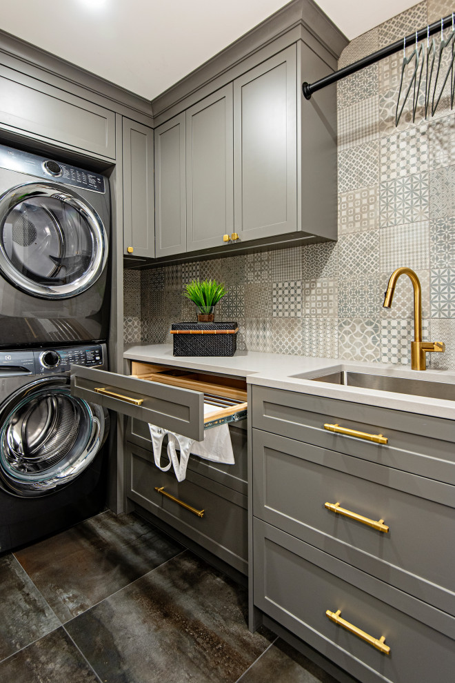 Design ideas for a small transitional l-shaped laundry room in Toronto with an undermount sink, shaker cabinets, grey cabinets, quartz benchtops, grey splashback, ceramic splashback, grey walls, ceramic floors, a stacked washer and dryer, black floor and grey benchtop.